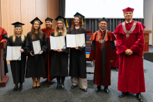 A group of graduates in black gowns is posing for a photo with their diplomas. To the right of them stand the dean and the vice-rector.