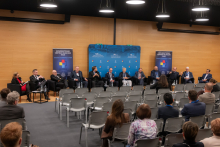 A discussion panel in a conference hall with twelve participants seated in black armchairs arranged along a wooden wall. In front of each armchair, there are small tables with water bottles and glasses. In the background, a blue backdrop is visible with inscriptions highlighting the anniversaries.