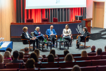 Five people are sitting on stage in armchairs and talking. In the background, other people can be seen watching those on stage.
