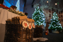 Baskets filled with packaged gingerbread are placed on the floor in a hall decorated for Christmas. In the background, Christmas trees adorned with lights are shining. Bright snowflake-shaped projections are visible on the wall.