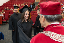 A young smiling graduate is receiving her diploma from the vice-rector. In the background, behind her, other graduates are visible waiting to receive their diplomas.