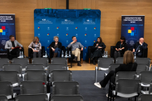 Seven people seated in black armchairs arranged in a row on stage in a conference hall. In front of each armchair, there are small tables with water bottles and glasses. In the background, a blue backdrop is visible with inscriptions highlighting the anniversaries.
