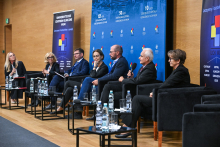 Seven people seated in black armchairs arranged in a row on stage in a conference hall. In front of each armchair, there are small tables with water bottles and glasses. One of the participants is holding a microphone, indicating that a discussion is taking place. In the background, a blue backdrop is visible with inscriptions highlighting the anniversaries.