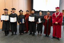 A group of graduates in black gowns is posing for a photo with their diplomas. To the right of them stand the dean and the vice-rector.