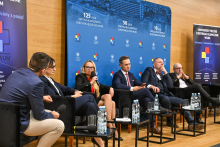 Six people seated in black armchairs arranged in a row on stage in a conference hall. In front of each armchair, there are small tables with water bottles and glasses. One of the participants is holding a microphone, indicating that a discussion is taking place.