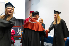 On the right, a young female graduate is receiving her diploma from the dean and the vice-rector. On the left, another graduate, with a big smile, is raising her diploma in a victorious gesture.