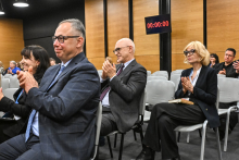 Several people seated in rows on chairs in a conference hall, facing the stage. They are dressed in elegant business attire and have their hands raised in a clapping gesture