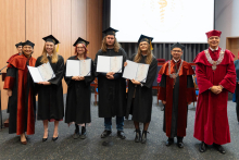 A group of graduates in black gowns is posing for a photo with their diplomas. To the right of them stand the dean and the vice-rector.