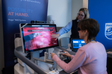 Two people are using a Medtronic medical simulator; one operates the controllers in front of a screen displaying an image of the inside of the body, while the other observes with a tablet. In the background, there are banners featuring the company’s slogans.