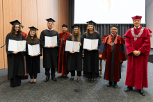 A group of graduates in black gowns is posing for a photo with their diplomas. To the right of them stand the dean and the vice-rector.