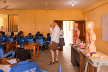 A classroom with students in blue shirts sitting at wooden desks, while the teacher stands next to the door. On the table, there are anatomical models representing internal organs.