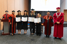 A group of graduates in black gowns is posing for a photo with their diplomas. To the right of them stand the dean and the vice-rector.