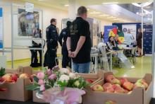 A police information booth in a modern building interior. In the background, officers and visitors are at tables with promotional materials, while in the foreground there are crates of apples and a bouquet of flowers.