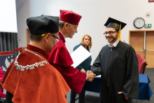 A young graduate is receiving a diploma from the vice-rector and the vice-dean. The graduate is shaking hands with the vice-rector.
