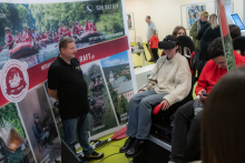 A booth promoting outdoor activities, including rafting and paintball. Next to a banner with information about rafting, a man is standing, while another person is sitting in an armchair wearing a VR headset. In the background, people can be seen observing and using their phones.