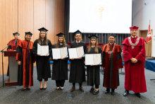 A group of graduates in black gowns is posing for a photo with their diplomas. To the right of them stand the dean and the vice-rector.