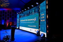 The stage during the Jubilee Gala of the University Clinical Center WUM, illuminated with blue and purple lights. In the background, a large screen displays the text: “From History to Innovation. Jubilee of the University Clinical Center WUM. November 27, 2025,” and on the right side, the inscription “UCK WUM Gala.” On the stage, there are microphones and floral decorations.