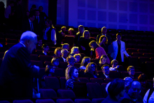 The auditorium audience in semi-darkness, with people sitting and standing, illuminated by blue and yellow lights.