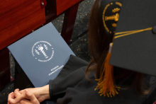 A young woman in a gown and a cap with a yellow tassel. She is sitting on a red chair, with her hands clasped on her lap. On her lap lies a navy-blue folder with the inscription “Warsaw Medical University, Faculty of Pharmacy.”