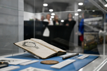 A display case featuring an open book, several documents, and a pen, accompanied by small plaques with descriptions of the exhibits. In the background, people can be vaguely seen conversing, likely within an exhibition or museum space.