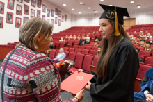 A young female graduate, dressed in a black gown and cap, is receiving a red folder with a distinction from an elegantly dressed woman, Dr. Aldona Wierzbicka-Rucińska.