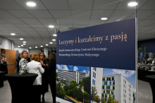 A poster with the text: “We Treat and Educate with Passion. Jubilee of the University Clinical Center of the Medical University of Warsaw,” along with two photos of modern buildings. In the background, people are standing by high tables during an event in a bright hall with ceiling lighting.