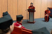 Prof. Grażyna Sygitowicz, dressed in a black and orange gown, is speaking from behind the lectern. Next to her, the dean is seated. In the foreground, other participants of the ceremony are visible.