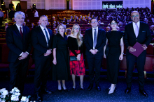 Seven people standing in a row in an auditorium with red seats, with event participants visible in the background. The individuals are dressed formally, indicating the ceremonial nature of the gathering.
