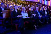 A large group of people seated in an auditorium during an event, with several individuals in the front row holding documents or devices. In the background, additional rows of participants can be seen, while lighting in shades of blue and warm tones creates a ceremonial atmosphere.