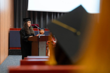 A young woman, dressed in a black academic gown, is standing behind the lectern and speaking.