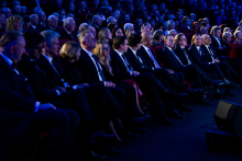 A large group of people seated in an auditorium during an event, with several individuals in the front row holding documents or devices. In the background, additional rows of participants are visible, while lighting in shades of blue and warm tones creates a ceremonial atmosphere.