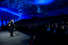 A ceremonial event in a large auditorium filled with participants. On the stage, three people are standing at microphones, presumably giving speeches. The lighting features blue tones with circular patterns on the ceiling, and portraits are visible on the walls.