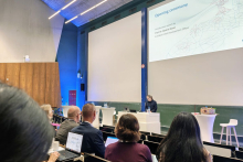 A lecture hall with a large screen displaying the slide “Opening ceremony.” At a white table on the podium stands a female speaker, while conference participants are seated in the rows, some of them using laptops.