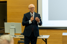 Professor Rafał Krenke, elegantly dressed, is standing in the middle of the hall and speaking. He is holding a microphone in his right hand, and behind him, part of a multimedia screen and a computer can be seen.