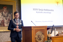 A man in academic attire with a rector’s chain stands at a lectern in a conference hall, against the backdrop of a screen displaying the text “35th Nobel Session of the Faculty of Medicine, Warsaw, December 15, 2025.” In the background, a portrait of a historical figure in ornate clothing is visible.