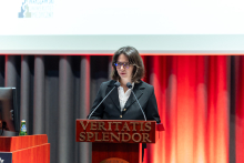A young, elegantly dressed woman is standing behind a lectern and speaking.