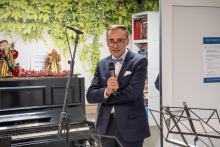 An elegantly dressed person is standing next to a piano decorated with Christmas ornaments and holding a microphone. In the background, there are green plants on the wall, a bookshelf, and music stands.