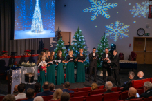 Nine people are standing in a row and singing, holding sheet music in their hands. Behind them, two Christmas trees decorated with lights are shining, and large snowflake projections can be seen on the wall. On the left side, there is a screen displaying an image of a bright Christmas tree in a winter forest. In the audience, a dozen or so people are seated, watching the performance. The hall is decorated with festive elements.