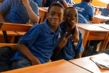 Two dark-skinned students in blue school uniforms are sitting next to each other at wooden desks, one of them making a “V” sign with their fingers.