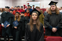 Graduates dressed in black academic gowns are standing in an auditorium among rows of red chairs. On the left, there is a group of people, also wearing gowns, entering the auditorium.