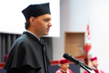 A young man, Szczepan Wąsik, wearing a black gown and cap, is standing behind the lectern and speaking. In the background, other people dressed in academic gowns are visible.
