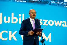 A person in a suit and tie is standing in front of a blue background with white lettering, holding a briefcase and speaking into a microphone, probably during a jubilee ceremony.