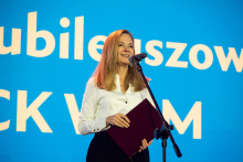 A woman in a white shirt is standing in front of a microphone, holding a briefcase and speaking. In the background, there is a blue backdrop with white lettering.