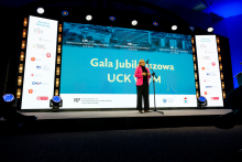A woman is standing on stage at a microphone, delivering a speech. In the background, there is a large screen displaying the text “Jubilee Gala UCK WUM” along with anniversary information: “125th Anniversary of the Dzieciątko Jezus Clinical Hospital,” “50th Anniversary of the Central Clinical Hospital,” and “10th Anniversary of the Postgraduate Medical Education Center.” On both sides, vertical banners with the institutions’ logos are visible.
