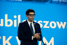 A man wearing glasses, a dark suit, a white shirt, and a tie is standing in front of a microphone with his hands clasped. In the background, there is a blue backdrop with white lettering.