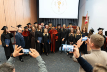 Fifteen people in gowns and caps standing in an auditorium. In the center, there are two people wearing red gowns. Several people are holding diplomas. In the foreground, two people are photographing the group with their phones.