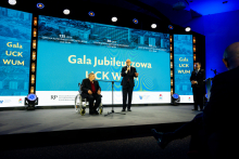 Three people are on stage during the “UCK WUM Jubilee Gala”; one person is seated in a wheelchair, another is speaking into a microphone, and the third is standing nearby.