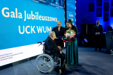 On stage at the “UCK WUM Jubilee Gala,” a man in a wheelchair is receiving a bouquet of flowers from two women.