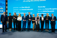 Eleven people in formal attire are standing on stage, each holding a small red box, likely containing an award. In the background, there is a large blue screen with the inscription “For Merits in Health Protection” and a list of names and academic titles.
