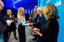 Kilka osób w stroju formalnym stoi na scenie, wymieniając uściski dłoni i odbierając małe czerwone pudełka, prawdopodobnie zawierające odznaczenia lub nagrody